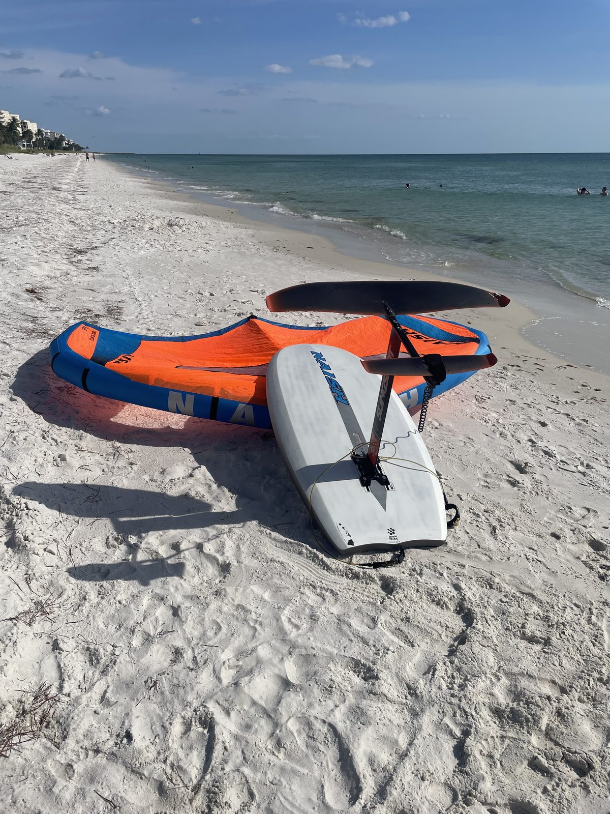 The used Naish wing, board, and hydrofoil setup, bought off Facebook Marketplace, laid out on a white sand beach with the Gulf in the background.